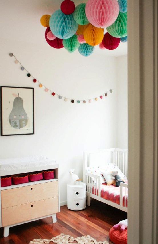 اتاق کودک با دیوار و تخت سفید و کفپوش چوبی که سقف آن با توپ های کاغذی رنگارنگ تزیین شده است
